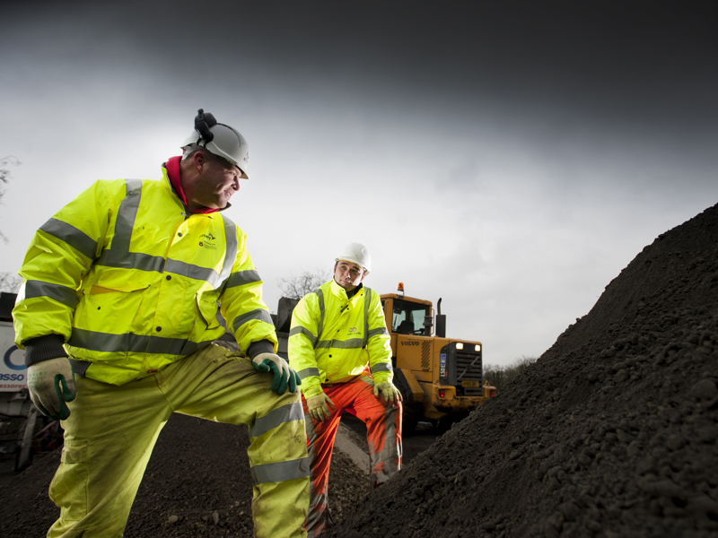 Two Amey male workers in PPE stood in front of a mound of earth.