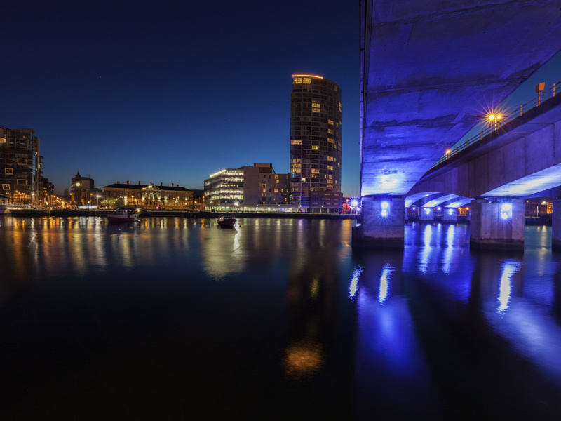 A night-time cityscape of Belfast, Ireland, featuring a lit bridge with blue lights reflecting on the calm water. A tall modern building and surrounding structures are illuminated in the background against a dark blue sky.