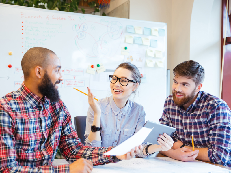 two men and a woman, sat at a desk in front of a whiteboard.