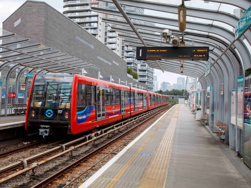 A red DLR train at Royal Victoria station, with a curved glass canopy and London skyline in the background