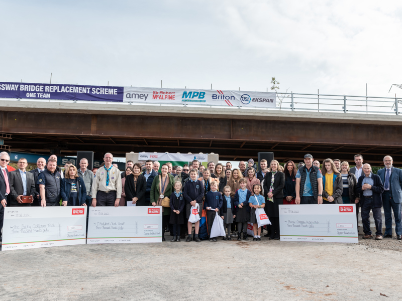 Image of a large crowd stood in front of a bridge, holding three cheques.
