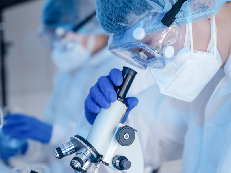 A person in PPE looking through a microscope.
