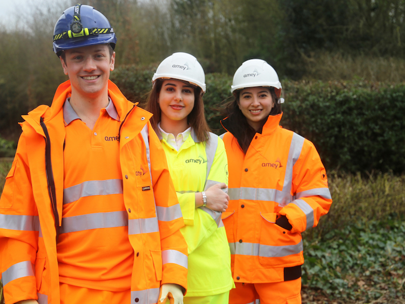 Three young Amey employees, stood one in front of the other, wearing PPE, smiling at the camera.