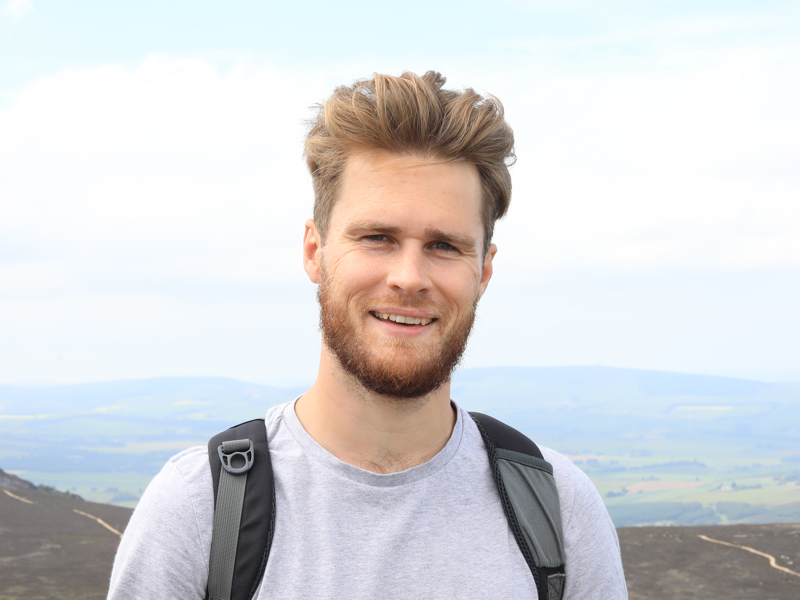 Head and shoulder image of a man stood on a mountain.