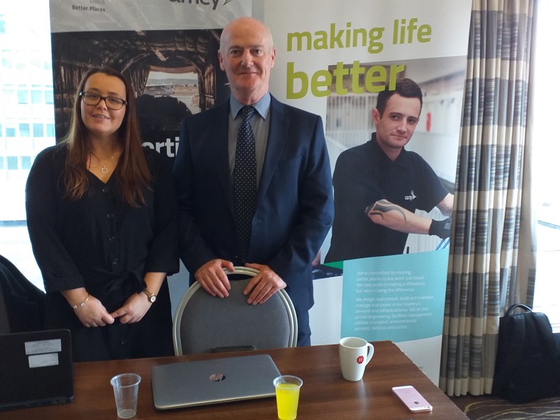 Image of Amey employees stood at a desk in front of Amey promotional banners.