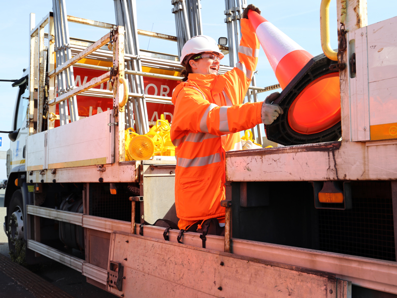 Female highway worker, in PPE taking an orange traffic cone off of a van.